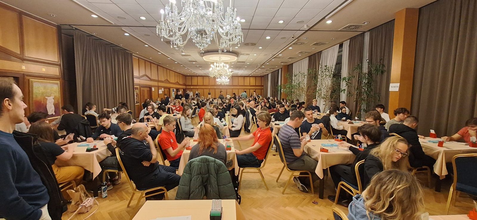 A full playing hall of young bridge players at tables under chandeliers